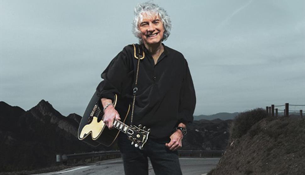Albert Lee wearing a black shirt and smiling at the camera, holding a guitar and standing on a road with mountainous terrain in the background