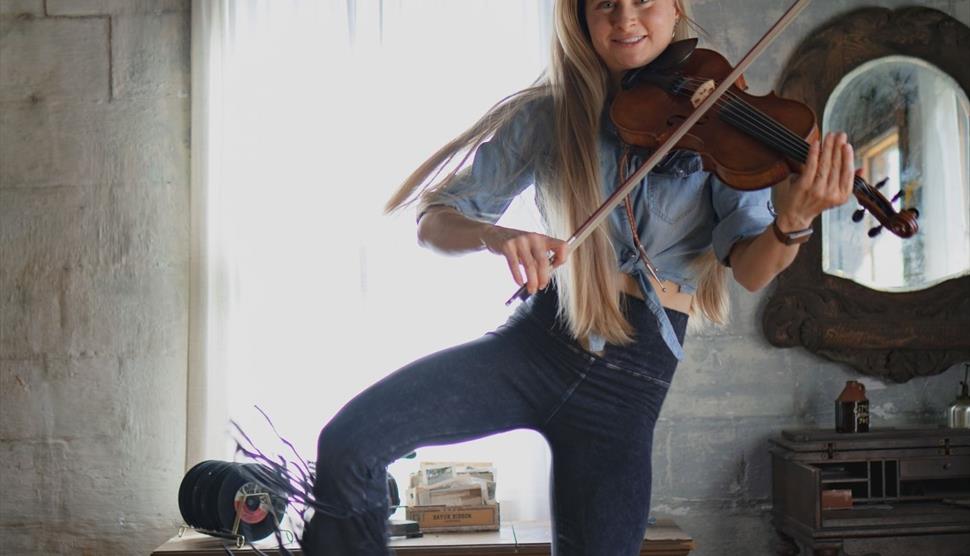 A woman plays the violin while jumping in mid-air inside a rustic room. Text promotes Hillary Klug's 2026 UK Tour on Saturday, July 11th in Aldershot