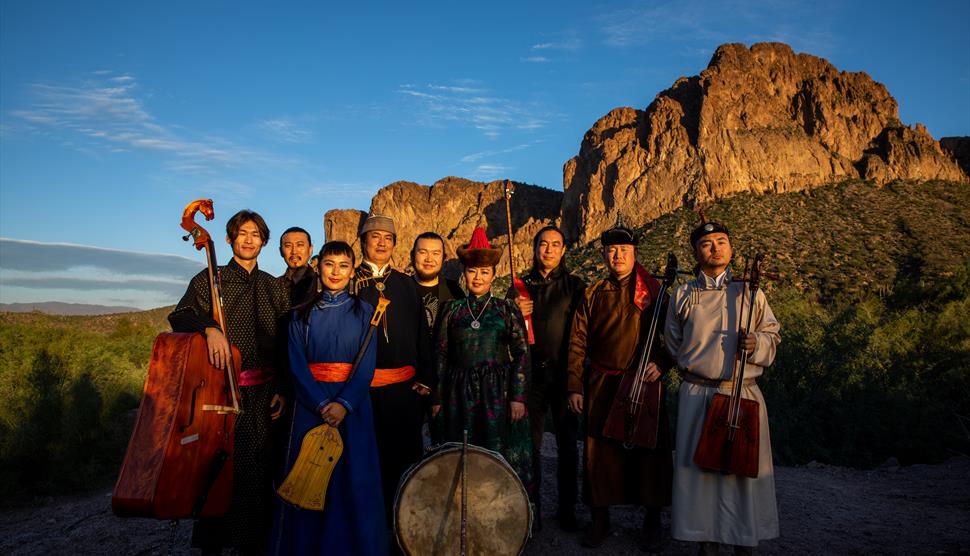 A group of musicians wearing traditional Mongolian clothes and stand in front of a hill. They're all holding traditional Mongolian instruments.