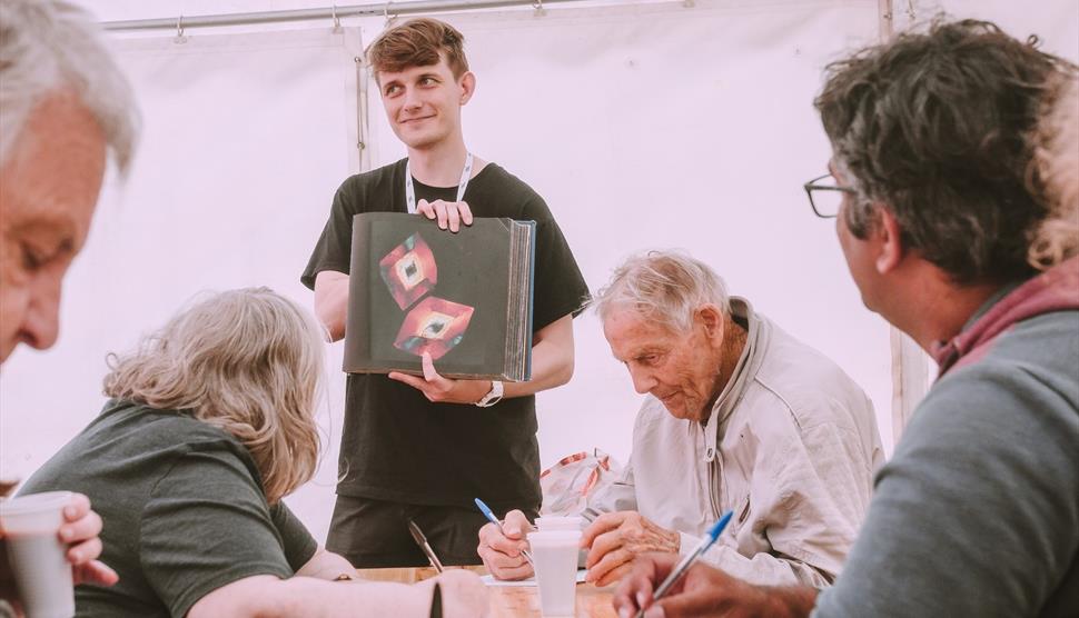 Poet delivering workshop, holding up a book, with participants sitting at a table.