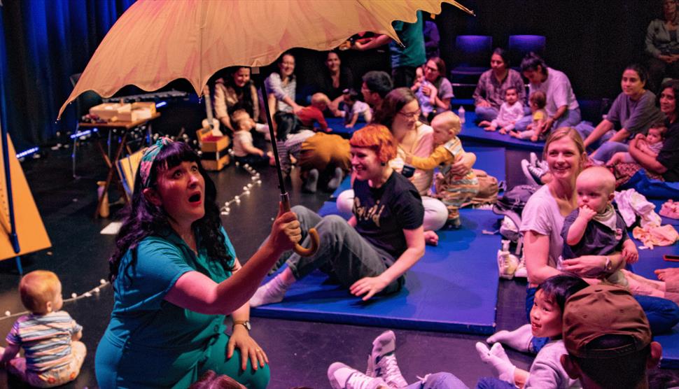 A woman with black hair is sat down and holding a yellow umbrella. Parents and babies are looking at her while sat on the floor.