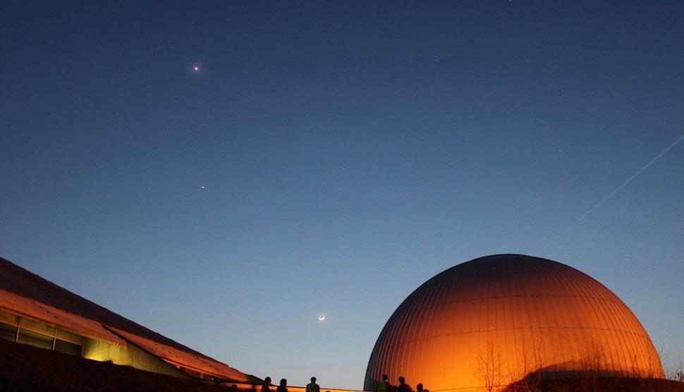 A dark sky with a few stars. The Science Centre and Planetarium are glowing orange against the night sky.