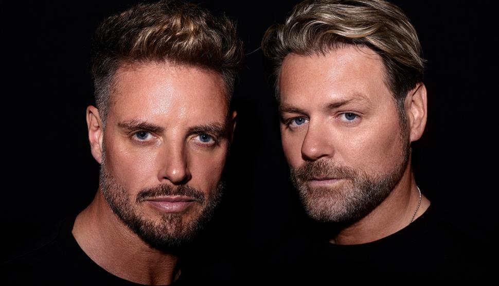 Keith Duffy and Brian McFadden in black tops with a black background looking at the camera