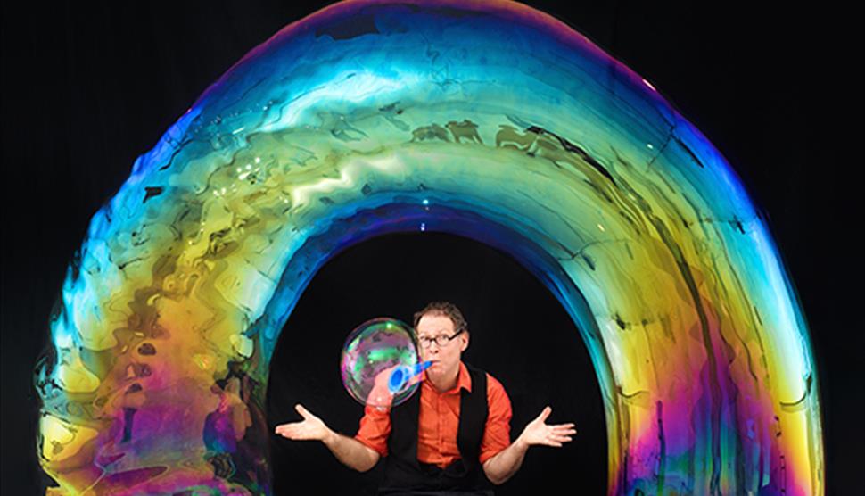 Louis Pearl (The Amazing Bubble Man) sitting underneath a bubble in the shape of a rainbow, with his hands outstretched. He is also blowing a bubble f