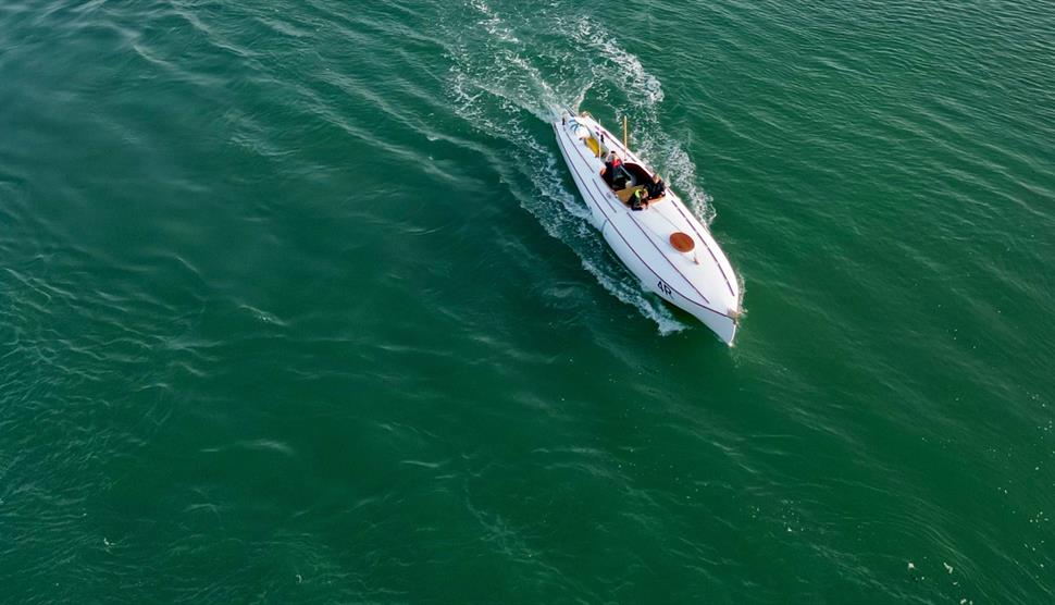Aerial view of a Coastal Motor Boat (CMB) speeding across the Solent, leaving a white wake behind as it travels over open water between coastline and