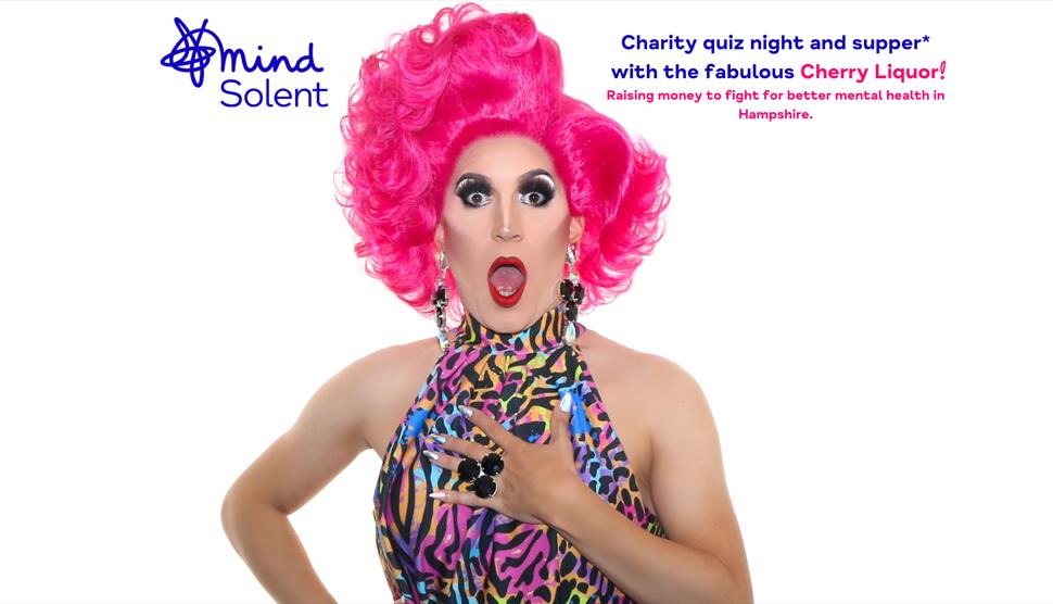 Drag artist Cherry Liquor is pulling a shocked face with their mouth open. They're wearing a big pink styled wig, dangling jewelled earrings and a mul