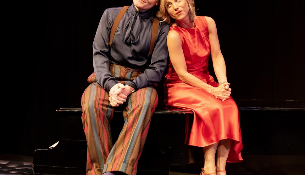 Liza Pulman and Joe Stilgoe are sitting together on top of a piano smiling.