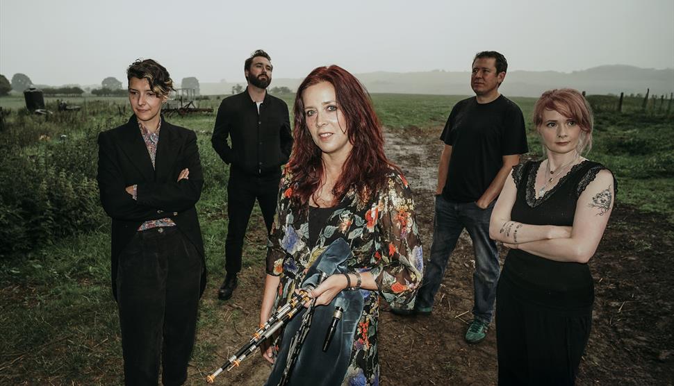 Five people in dark clothing stand on a muddy path in the countryside. A woman with red hair and a dark floral jacket stands in the centre.