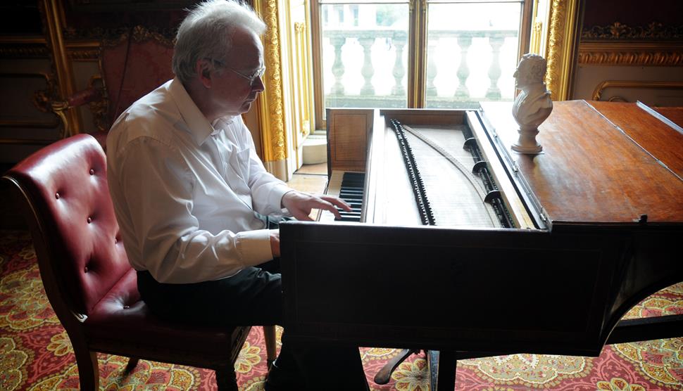 The image shows David Owen Norris sat on a red chair playing a piano by the window.