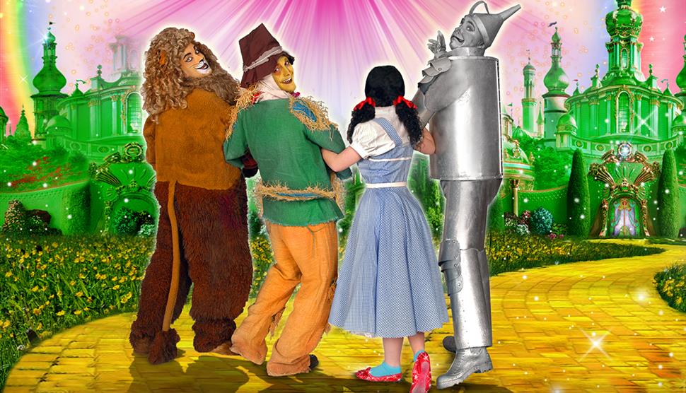 Four actors dressed as the Cowardly Lion, Scarecrow, Dorothy, and the Tin Man from The Wizard of Oz. They are all linking arms with the Cowardly Lion,