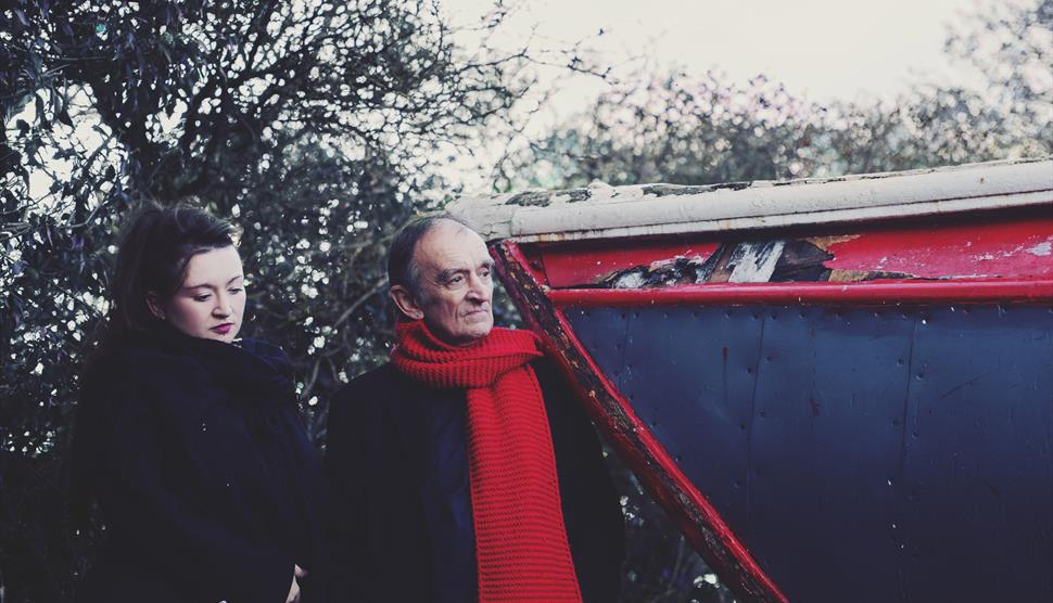 A woman with dark hair and a black coat stands next to an older man wearing a red scarf.