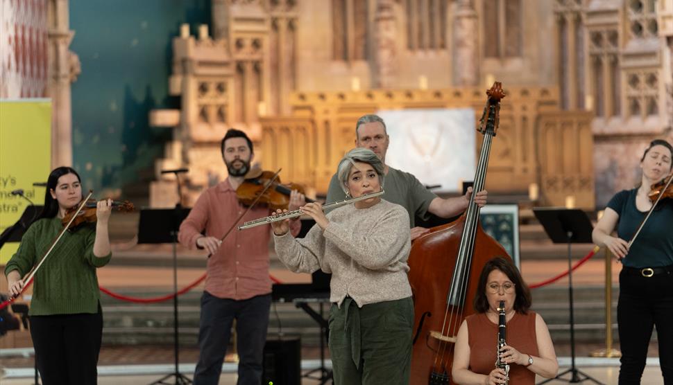 A group of classical musicians play their instruments. A woman with short, grey hair plays the flute in the centre, with several violinists and a doub
