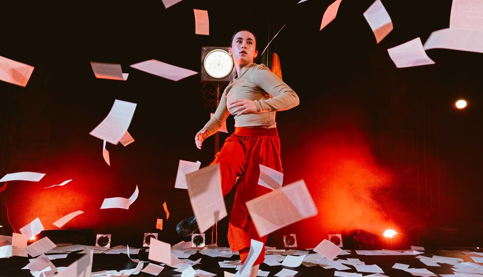 Circus performer stood centre stage with pieces of paper all around, flying up in the air