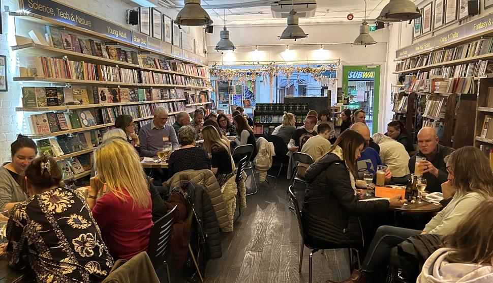 Groups of people sitting at tables in a bookshop with drinks taking part in a quiz