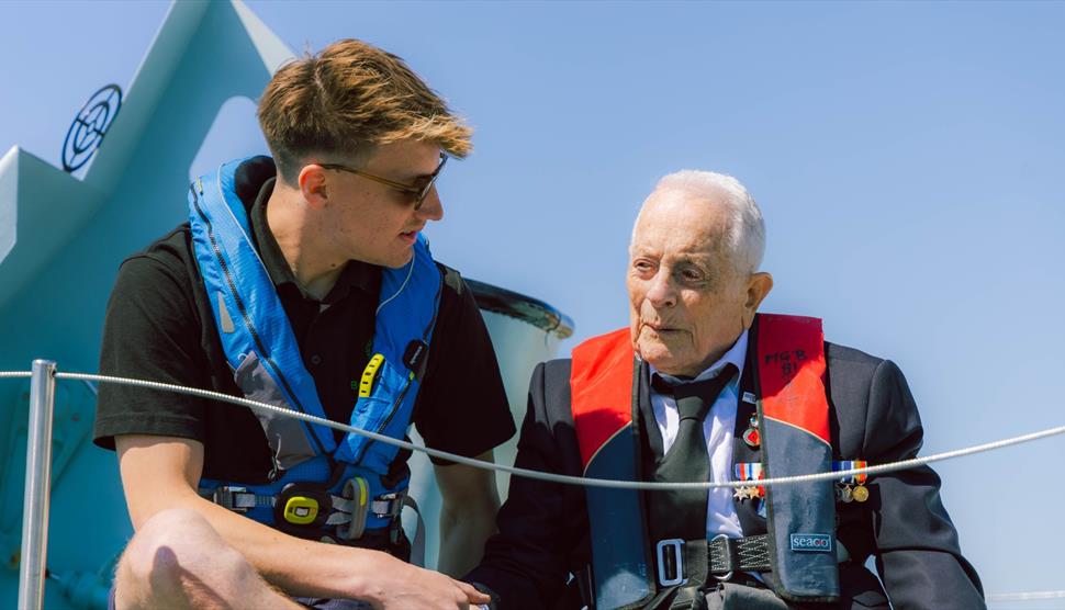 PHQ boat team member sitting and talking with a WWII veteran aboard Motor Gun Boat 81, sharing a moment of conversation on the historic vessel.