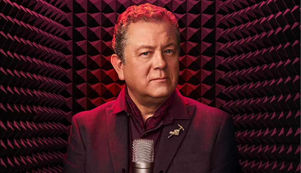 Jon Culshaw wearing a dark suit with a patterned shirt and a pin on his lapel. He's standing in front of a professional microphone soundproofing foam