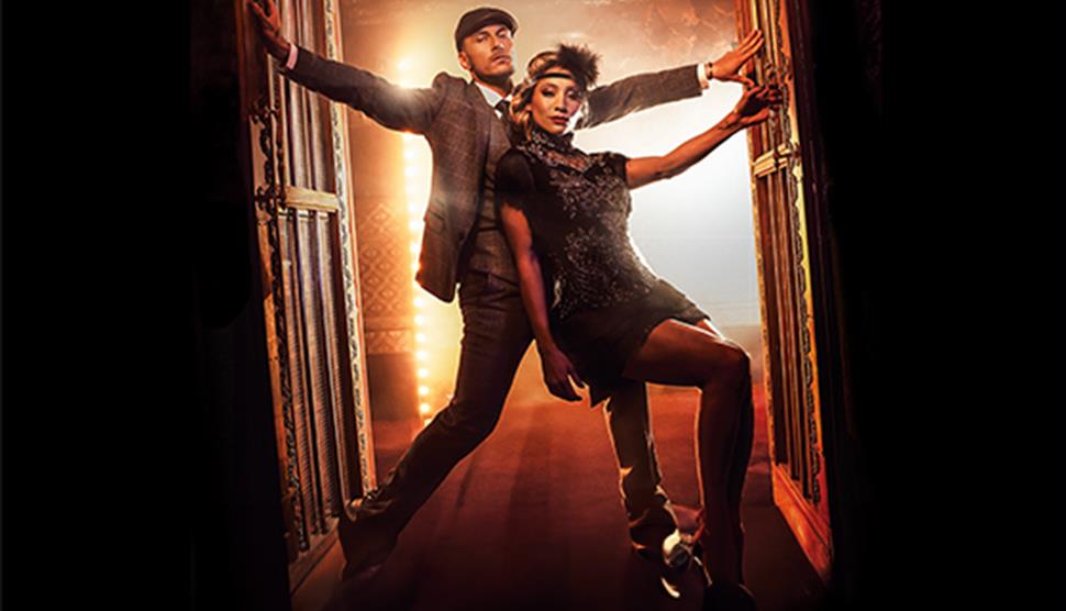 Gorka Marquez and Karen Hauer stood  in a doorway with red/orange light behind them and black in front. Gorka is in a three piece grey suit with a fla