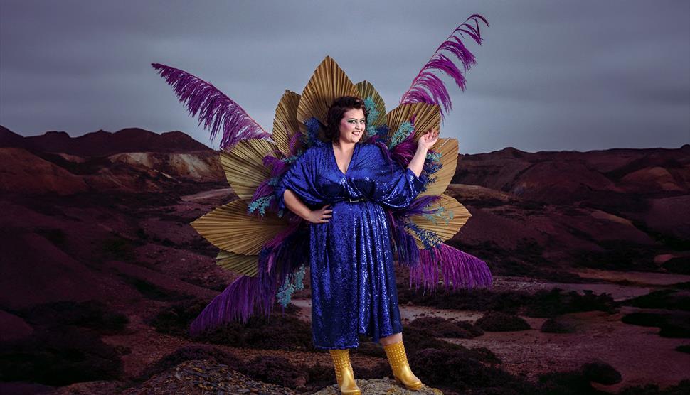 Kiri Pritchard-McLean stands in a sparkly blue dress on a rock, with a backdrop of a gloomy moor. Attached to her back is a large display of feathers,