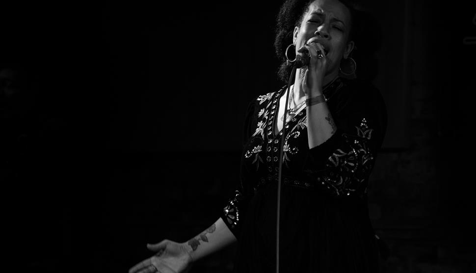 A black-and-white image of a woman singing into a microphone. She has large hoop earrings and a black top with embroidered leaves.