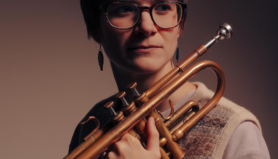 A woman with short hair glasses holds a trumpet.