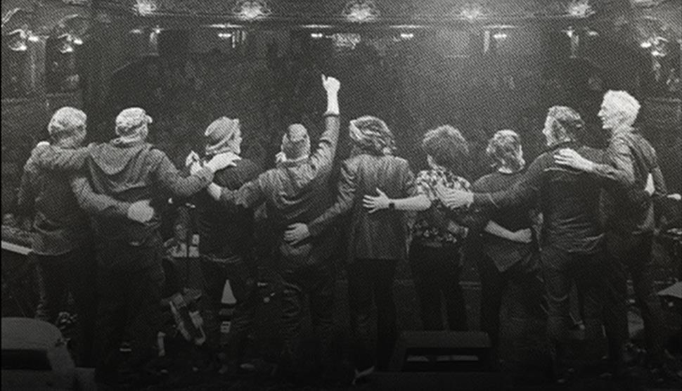 Black and white photo of the backs of nine members of the Levellers on stage, looking towards the audience with their arms around each other