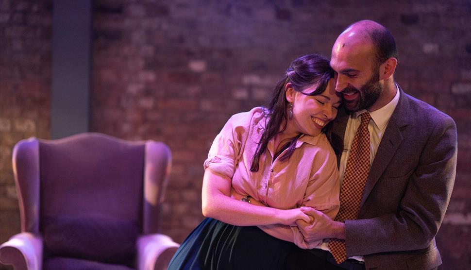 Two actors on stage, man is holding woman in an embrace. They are both smiling, they look in love. There is an empty chair behind them