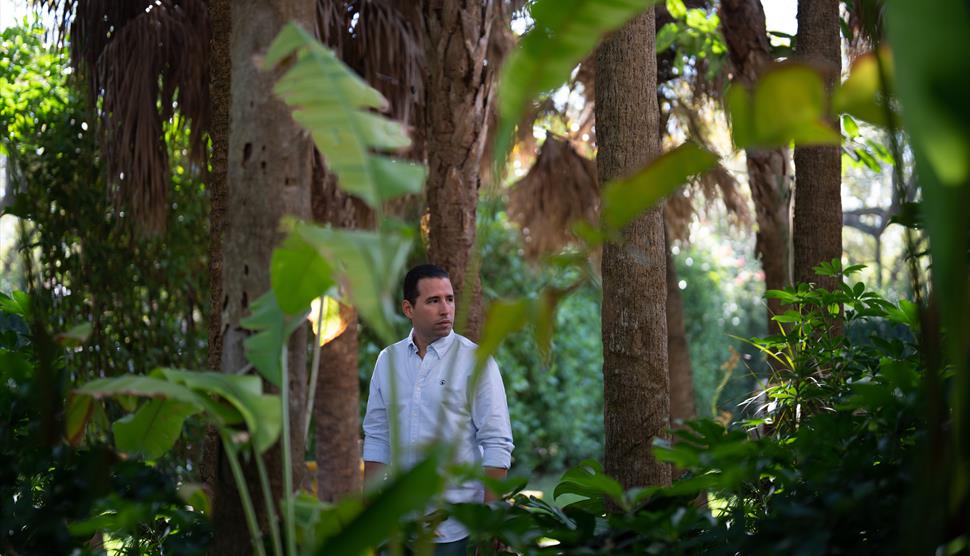 Alfredo Rodriguez is looking to the distance surrounded by palm trees and shrubbery.