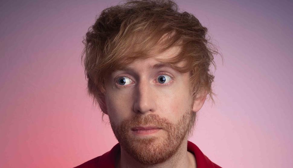 A close up of Mark Simmons wearing a shirt. He has a stubbly ginger beard and hair and wide, bright blue eyes