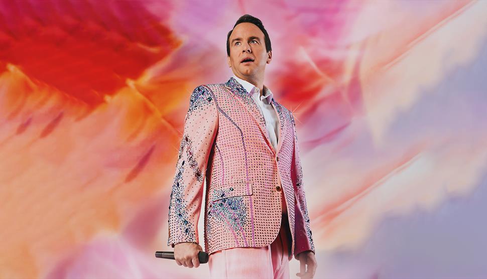 A man in a sparkly pink suit - Matt Forde - holds a microphone, standing against a vibrant, swirling pink and red background.