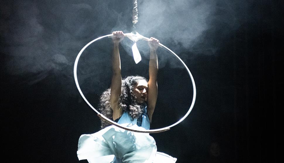 A circus performer dressed in a white dress hangs from an ariel hoop