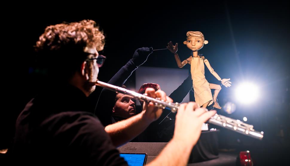 A flute player faces away from the camera whilst playing their instrument in front of a puppet being controlled by a puppeteer.