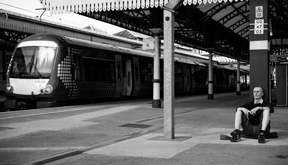 Hillview Art - man sat on floor of railway station with train in the background.