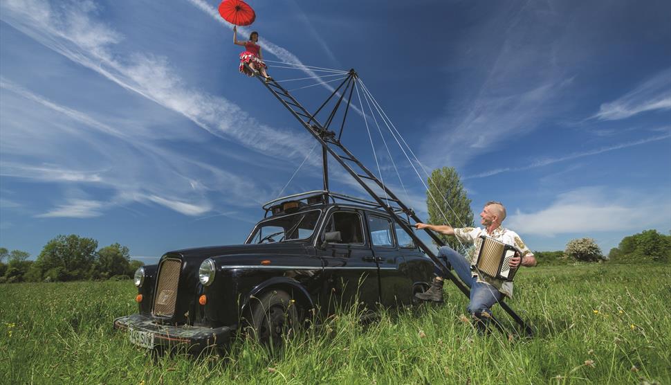 A black taxi in a field. A ladder is to the right hand side. A male stands next to it whilst a female hangs off the end.