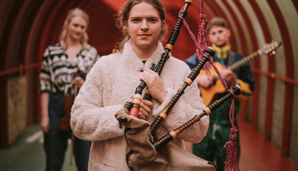 A man with long, brown hair and a white coat carries a bagpipe.