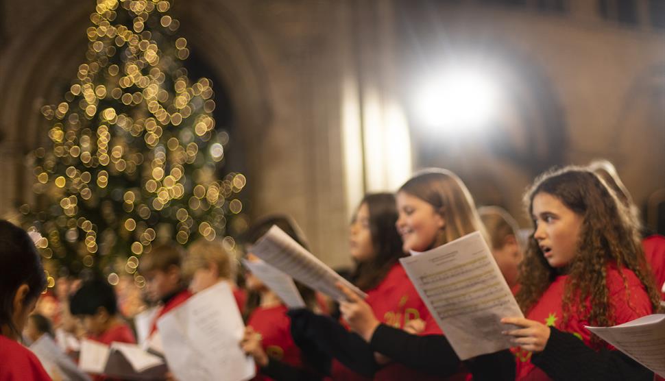 A group of children wearing red shirts sing from sheet music during a Gabrieli Roar concert performance in 2022. They stand in a warmly lit cathedral,