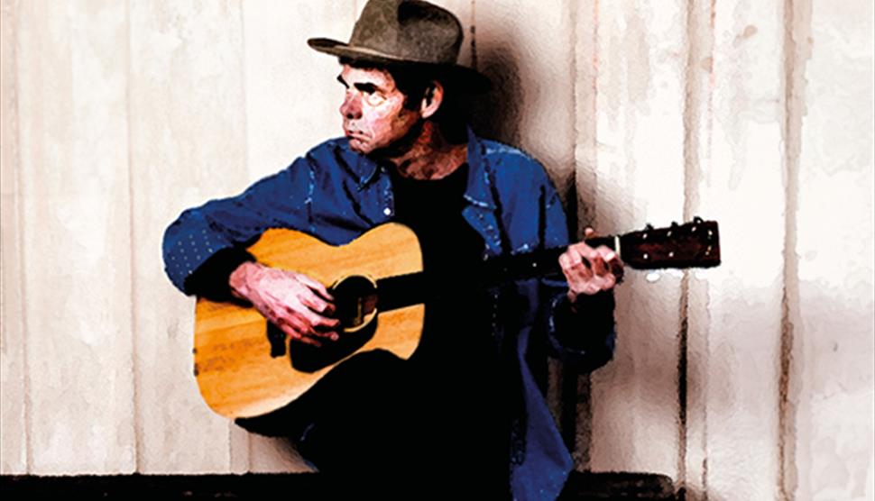 Rich Hall sitting down, playing an acoustic guitar. He is wearing a blue shirt and brown hat, looking away from the camera