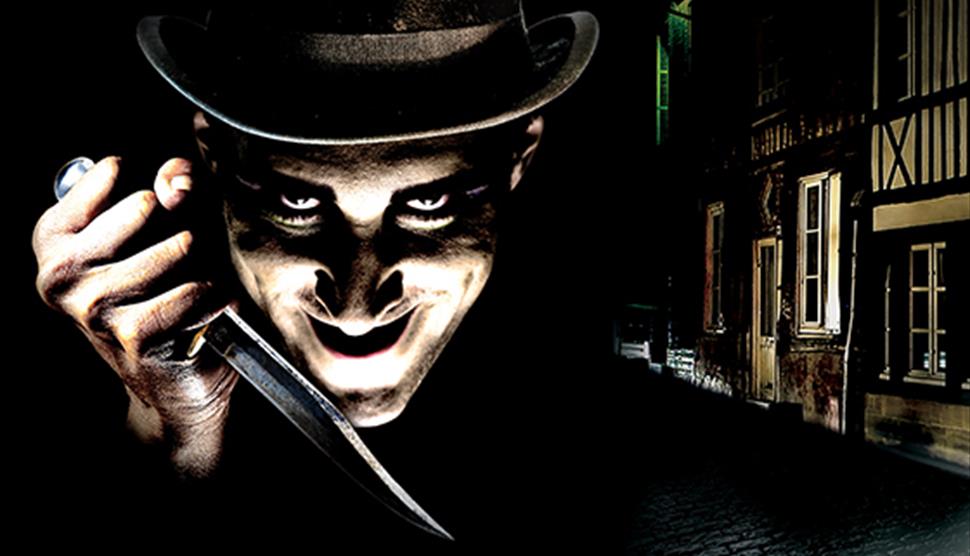 A very dark image with a Victorian era street in the background. The foreground is a man brightly lit from underneath, wearing a top hat and a knife l