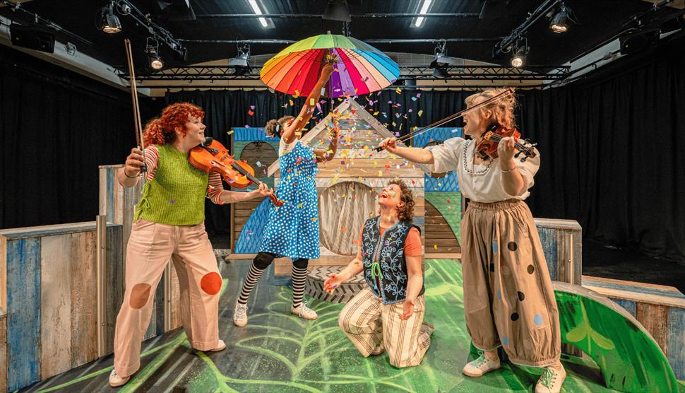 A woman wearing a checked blue and white dress holds up a rainbow umbrella. Confetti falls from the umbrella as two women holding violins smile.