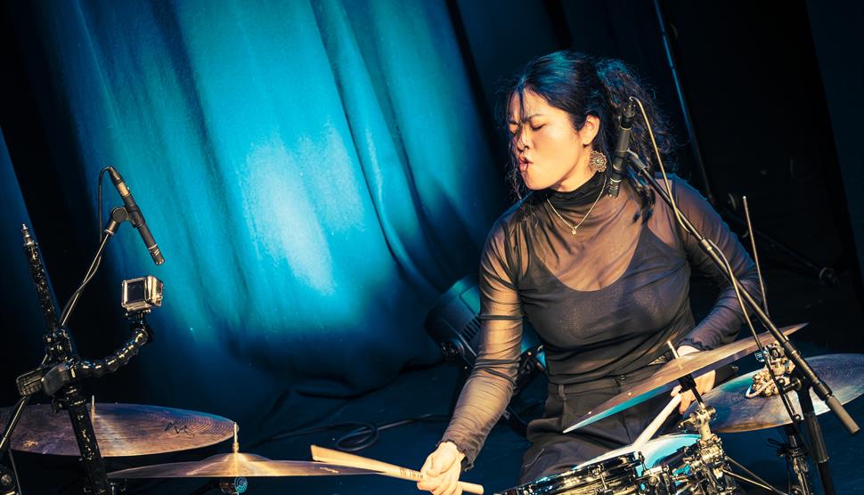 Sun-Mi Hong is sat in front of blue curtains and behind a drum kit and appears to be passionately drumming with her eyes closed.