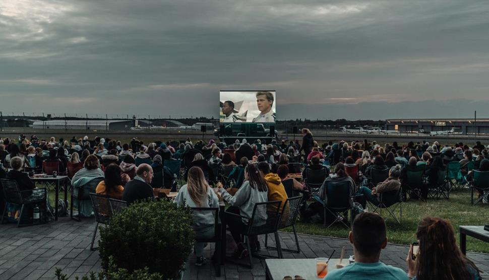 Outdoor movie night with viewers facing a large screen, airport visible behind screen