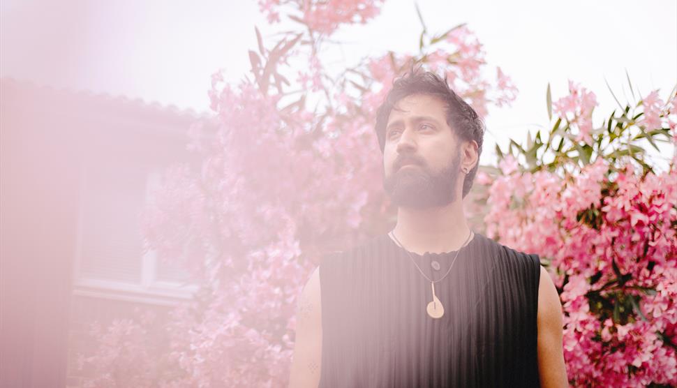 A man with dark hair and a black jumper stands in front of a cherry blossom tree.