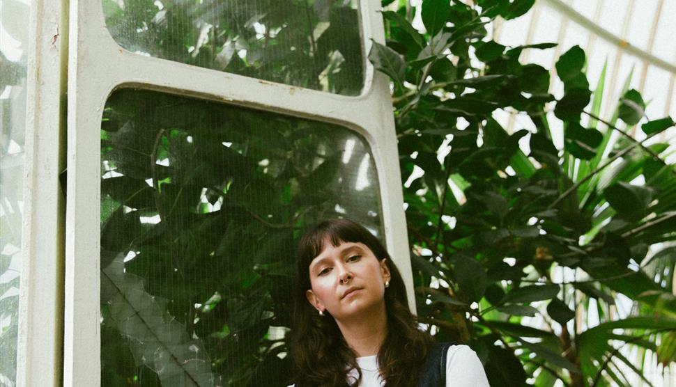 A woman with dark brown hair stands in front of dark green leaves in a forest.