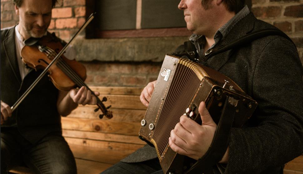 Two men are sat on a wooden bench. One man is playing the fiddle and the other is playing the accordion.