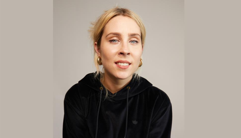 Poster image for Stevie Martin: Clout, showing the comedian in a black hoodie and stood against a brown background