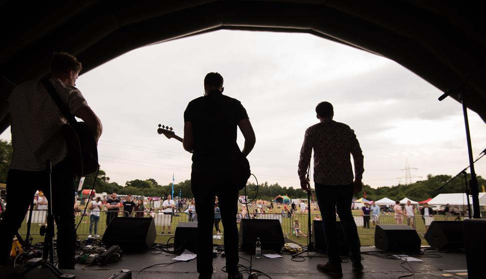 Image shows a band standing on an outdoor stage, seen from behind as they face a crowd at a festival. Three musicians with guitars are silhouetted aga