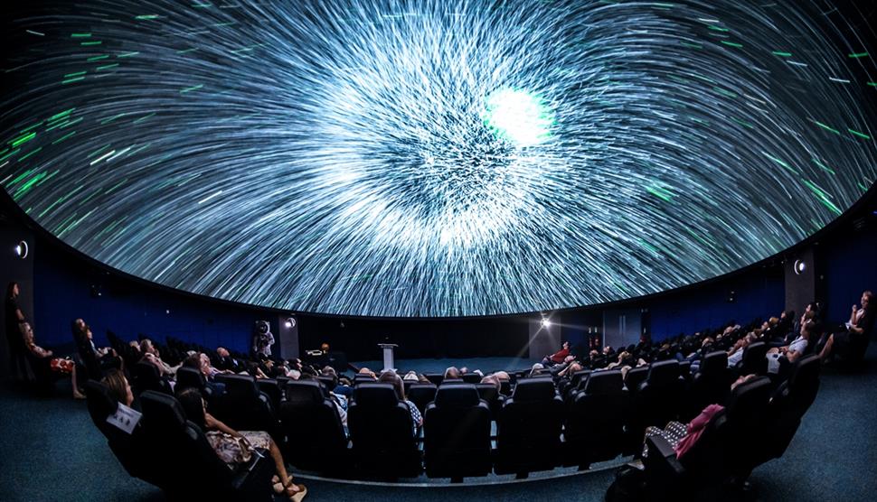 A blast of white light on a Planetarium dome. There are people sat in seats watching the screen.