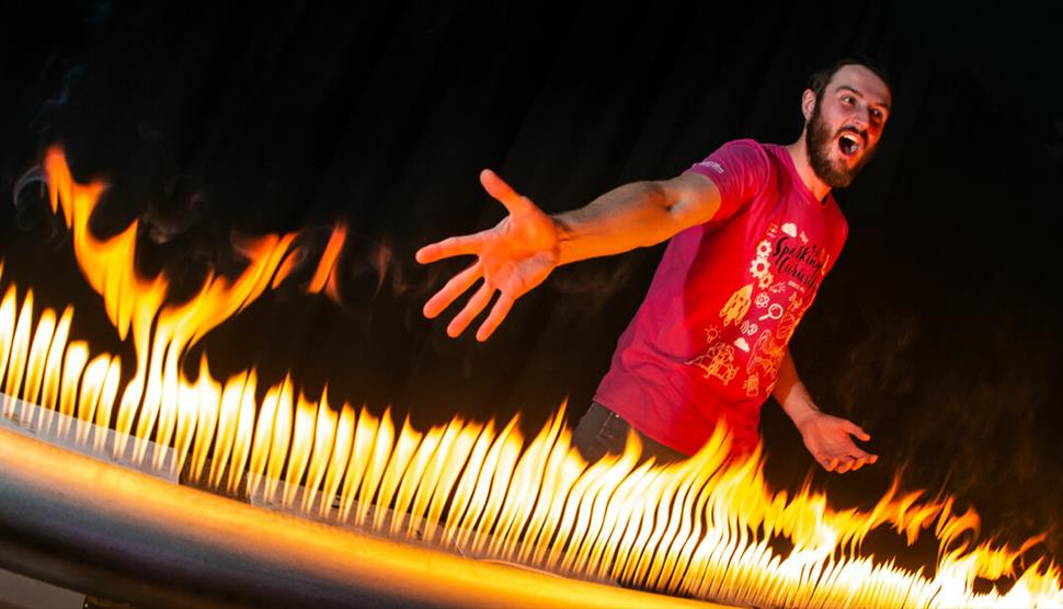 A man with a pink T-shirt on is standing in front of a large metal tube that has lots of holes in it. There is fire coming out of the holes and the ma