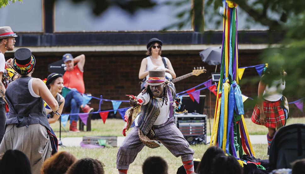 Male pointing into a crowd next to a maypole with colourful bunting and music being played in the background.