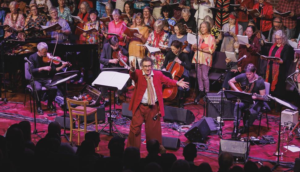 Harvey Brough reaches his arm out and smiles towards the camera. A choir and multiple instrumentalists are behind him.