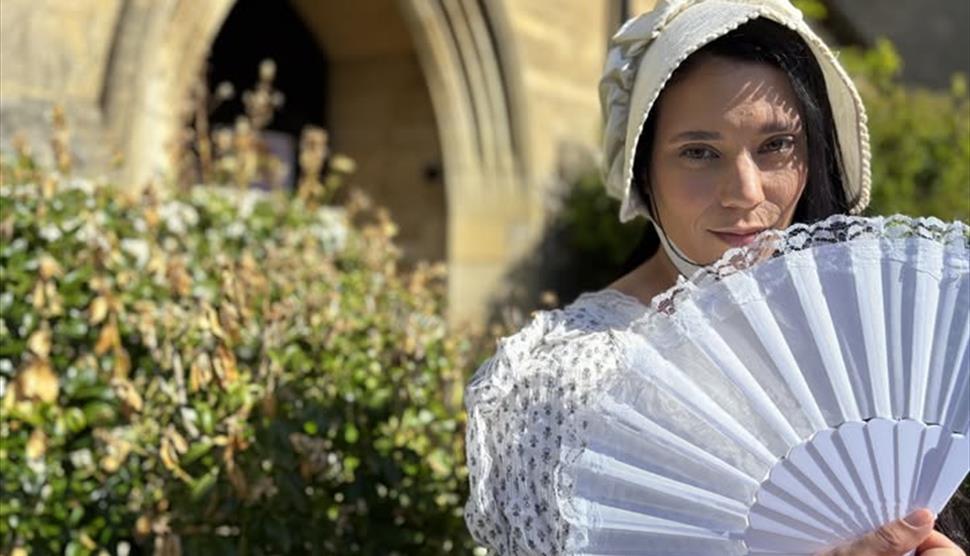 a woman dressed in white holding a fan standing in front of bushes